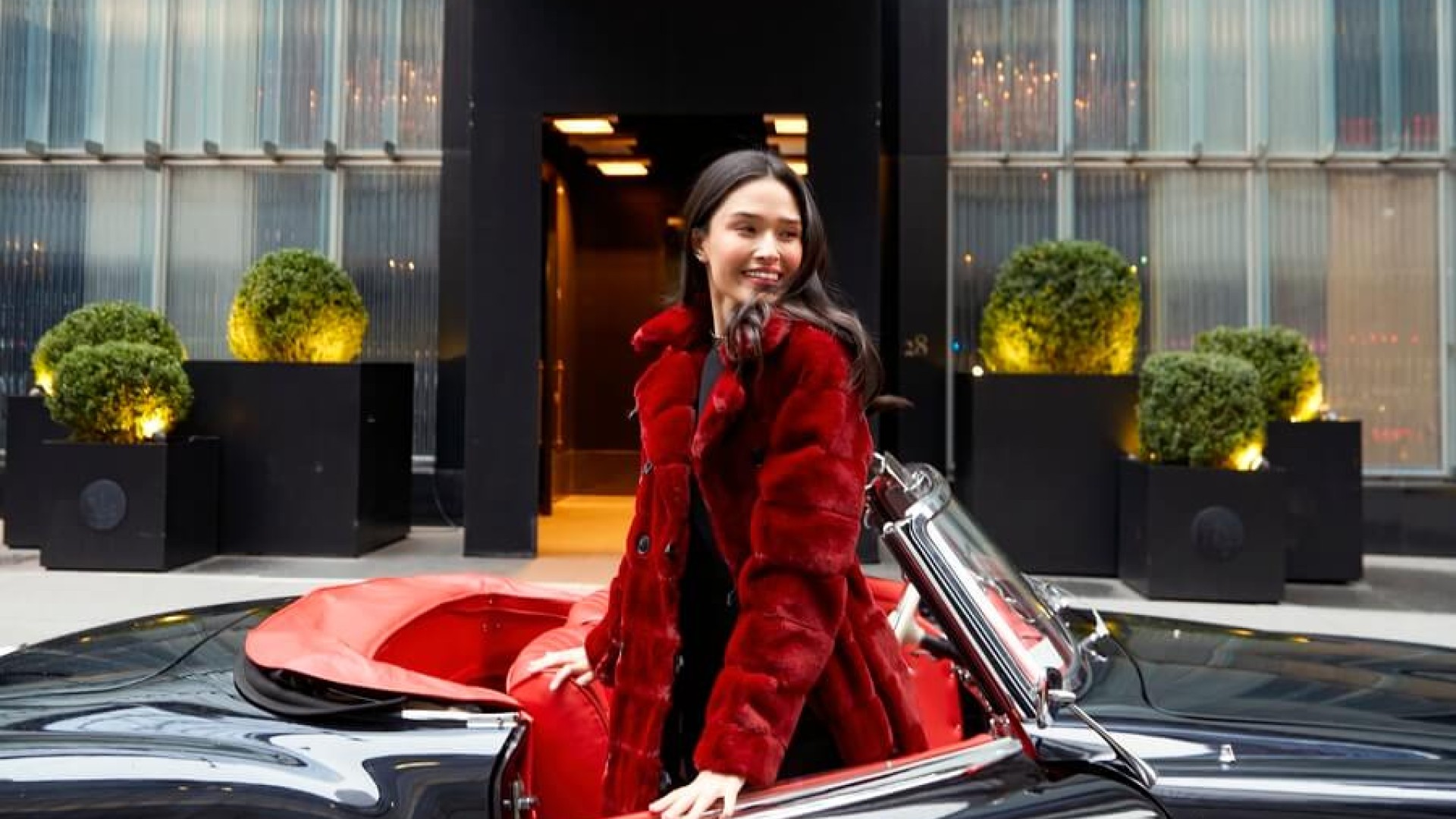 A woman wearing a red coat is seated in a classic car