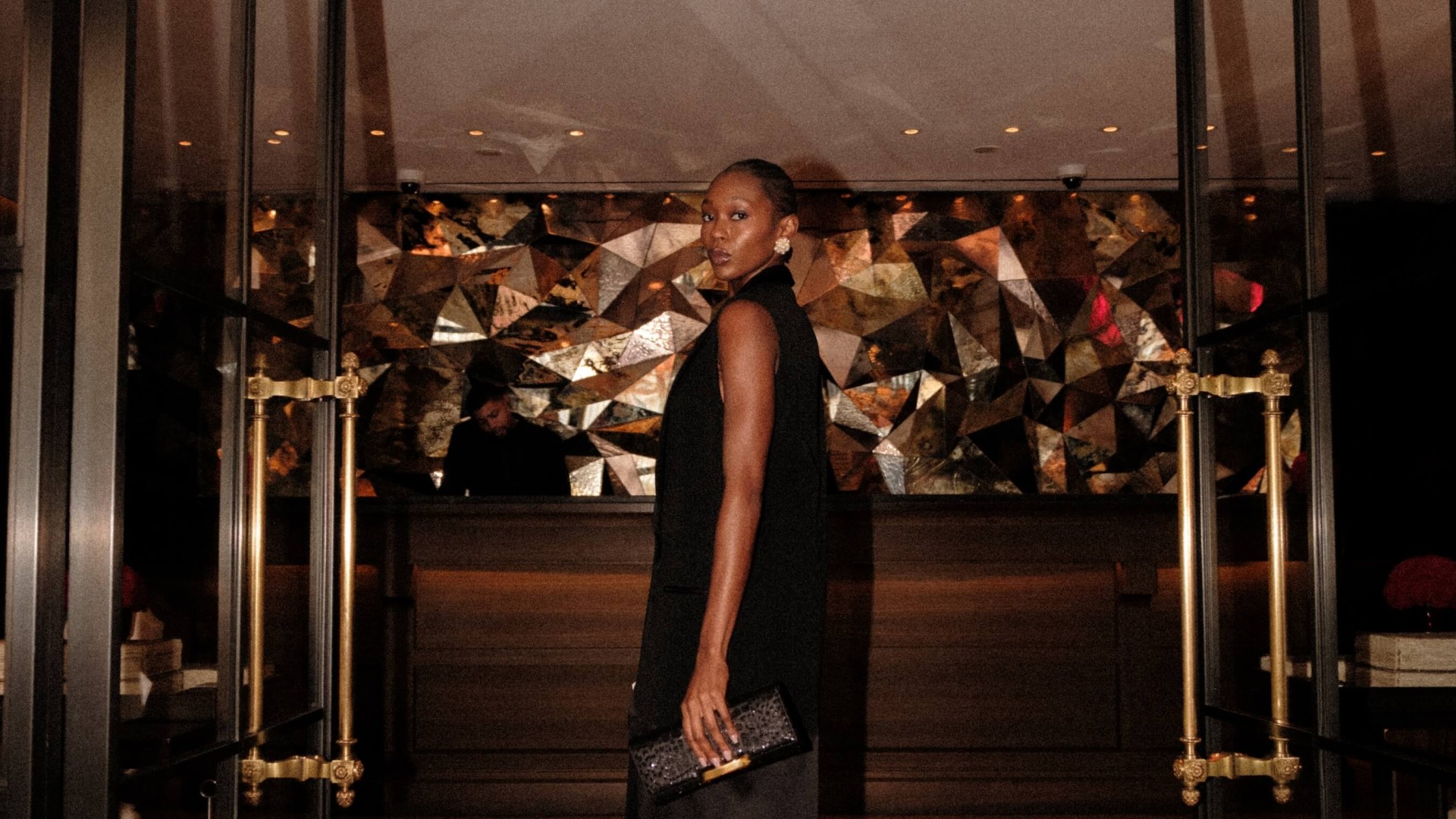 A woman in a black dress stands elegantly in front of a glass door