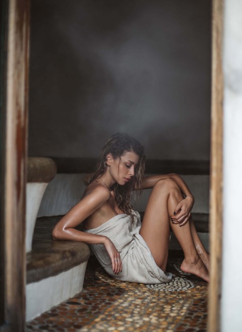 A woman wrapped in a towel sits on a tiled floor, appearing relaxed and contemplative