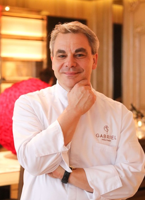 A man in a white chef's coat stands confidently in front of a table