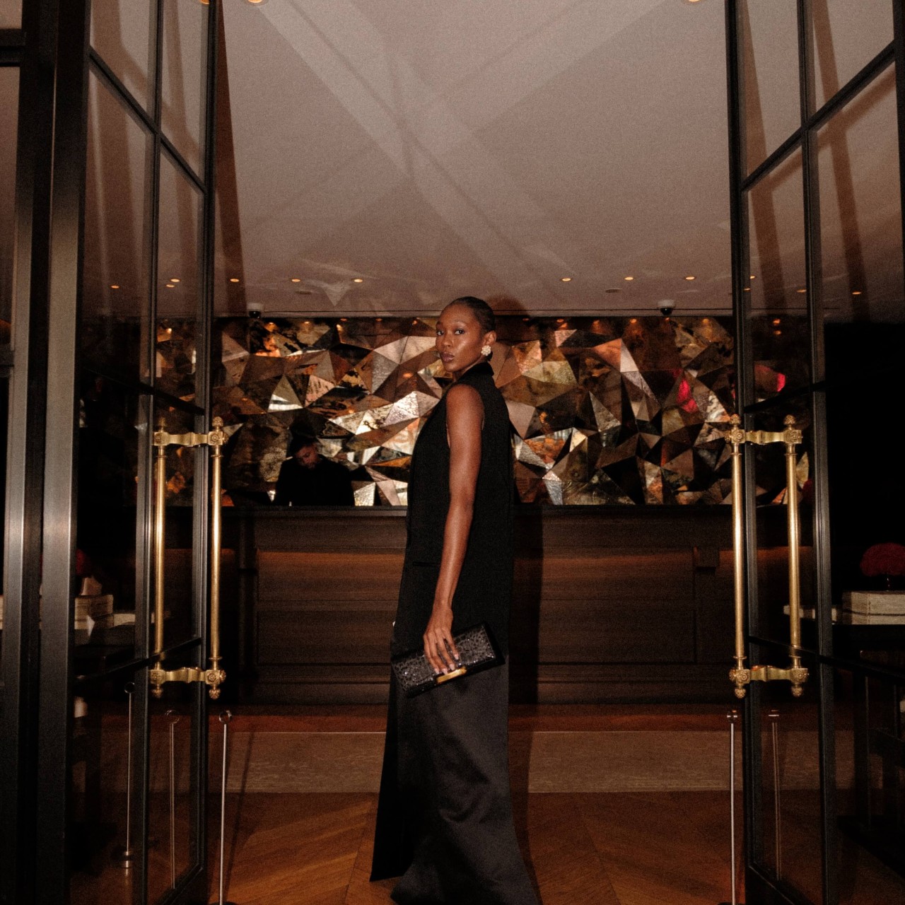 A woman in a black dress stands elegantly in front of a glass door