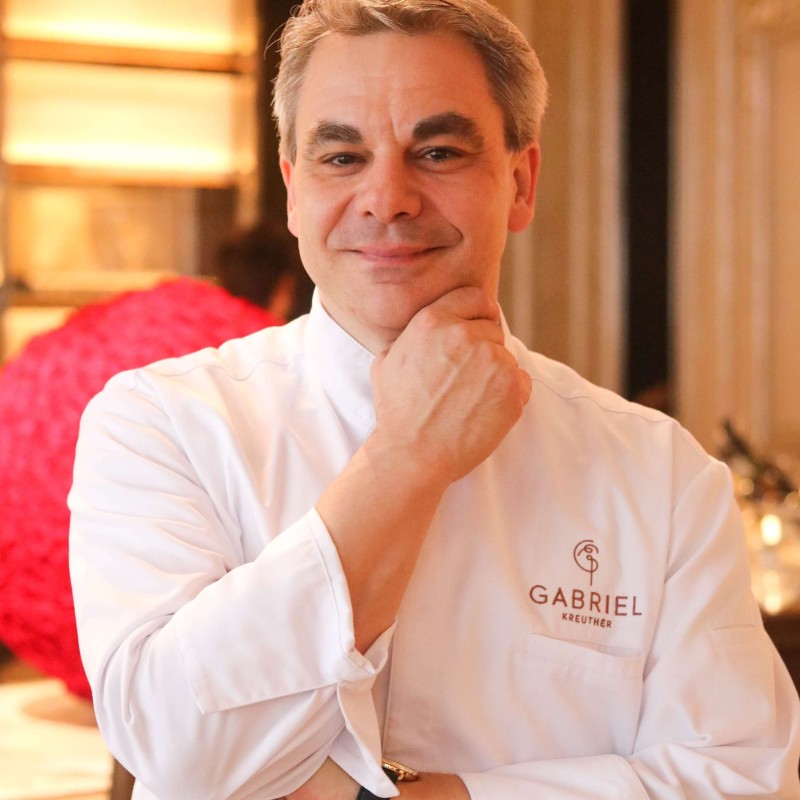 A man in a white chef's coat stands confidently in front of a table