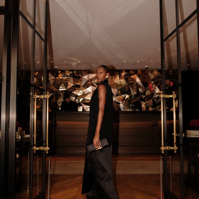 A woman in a black dress stands confidently in front of a large glass door