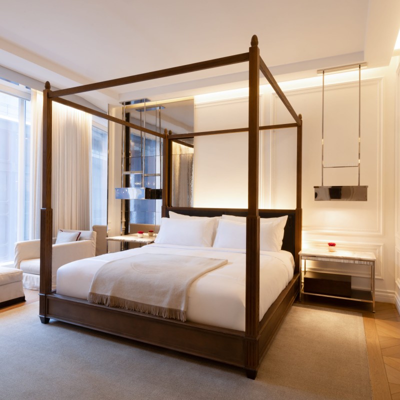 A large bed with a canopy in a bright guest room with floor to ceiling windows and a lounge area 
