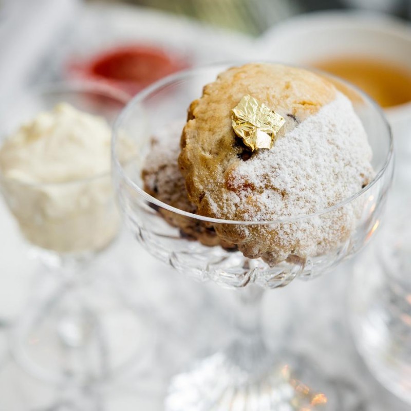 A layered dessert in a glass beside a steaming cup of tea on a wooden table.