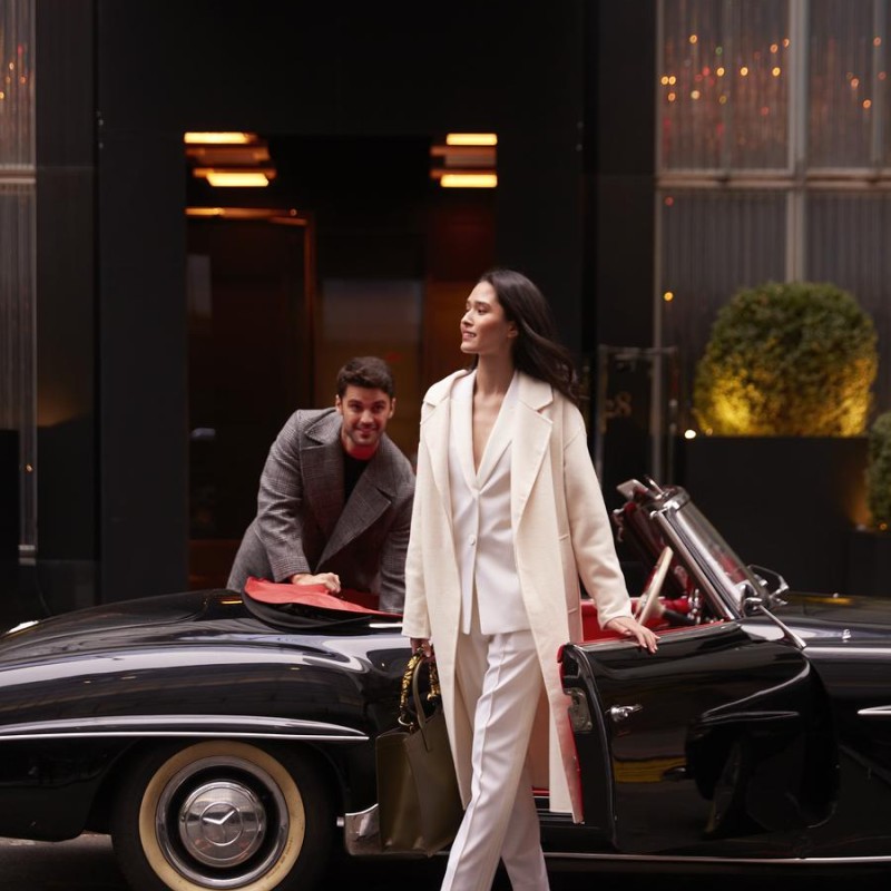 A couple stands next to a classic car, smiling and dressed in fashionable attire, highlighting the vehicle's retro appeal.